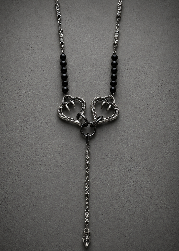 A gothic-style silver pendant necklace featuring two interlocking open hearts with sharp talons, flanked by black beads on a textured chain, set against a dark gray background.