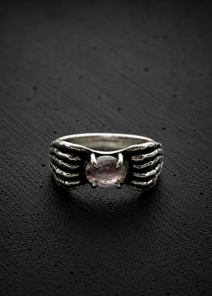 A silver ring featuring a textured winged design on the band and a central round pink gemstone, displayed on a dark textured background.