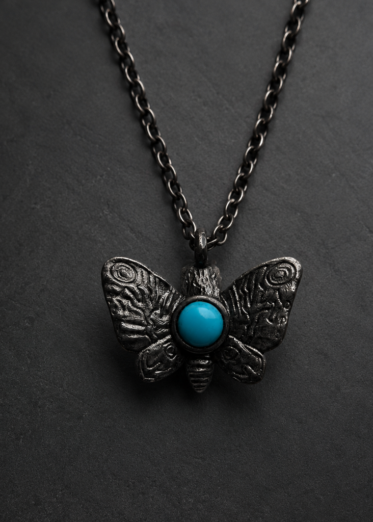 A dark metal moth pendant necklace with a vibrant turquoise blue stone in the center, displayed on a dark textured background.