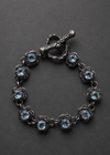 A dark silver-tone bracelet featuring nine round-cut light blue gemstones in a floral setting, connected by delicate chain links and finished with a decorative toggle clasp, displayed on a dark gray background.
