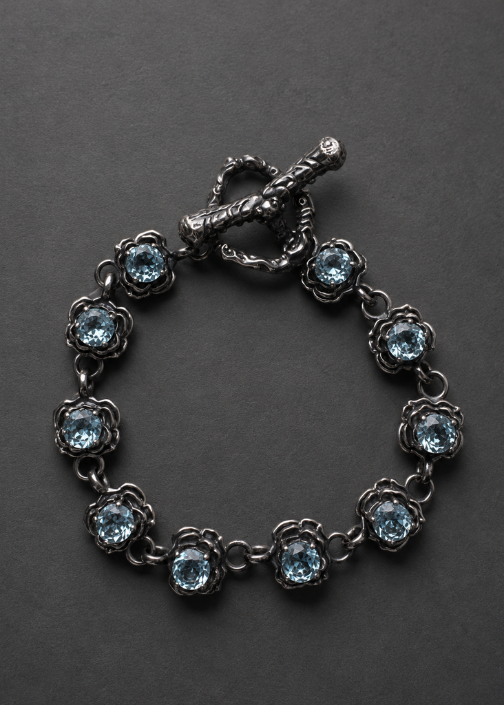 A dark silver-tone bracelet featuring nine round-cut light blue gemstones in a floral setting, connected by delicate chain links and finished with a decorative toggle clasp, displayed on a dark gray background.