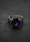 A silver ring with an ornate filigree band and a large, cushion-cut purple amethyst gemstone, displayed on a dark textured surface.
