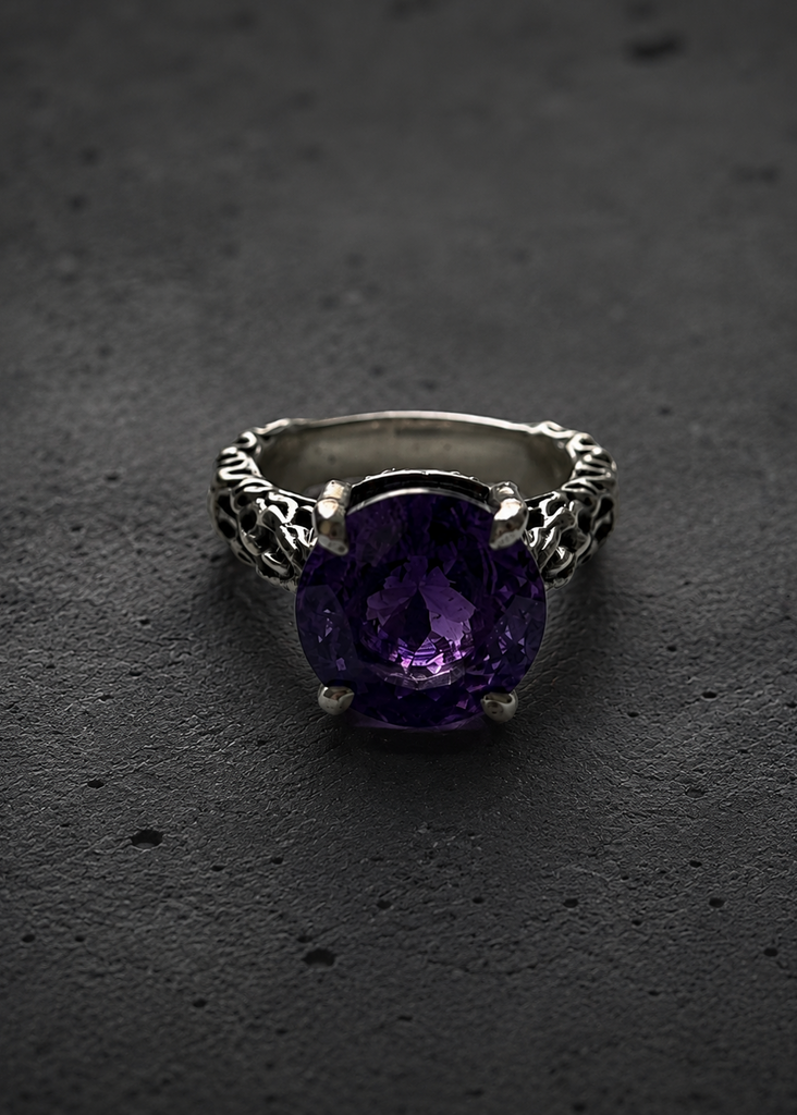 A silver ring with an ornate filigree band and a large, cushion-cut purple amethyst gemstone, displayed on a dark textured surface.