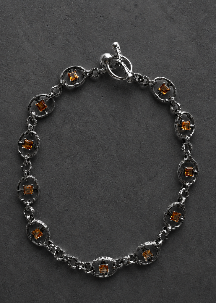 A silver-toned chain bracelet with oval links, each featuring a small amber-orange gemstone, displayed on a dark textured background.